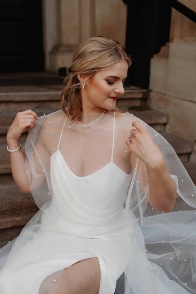 eine sitzende Braut trägt ein drapiertes Brautoberteil und eine Cape aus Perlen - Brautatelier Par Amour Hamburg